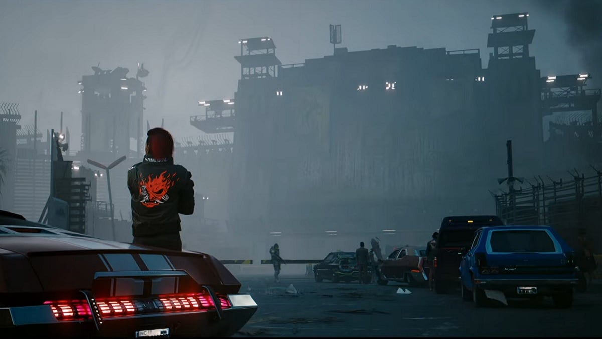 Cyberpunk 2077: V leaning on a car with a smoldering factory in the distance.