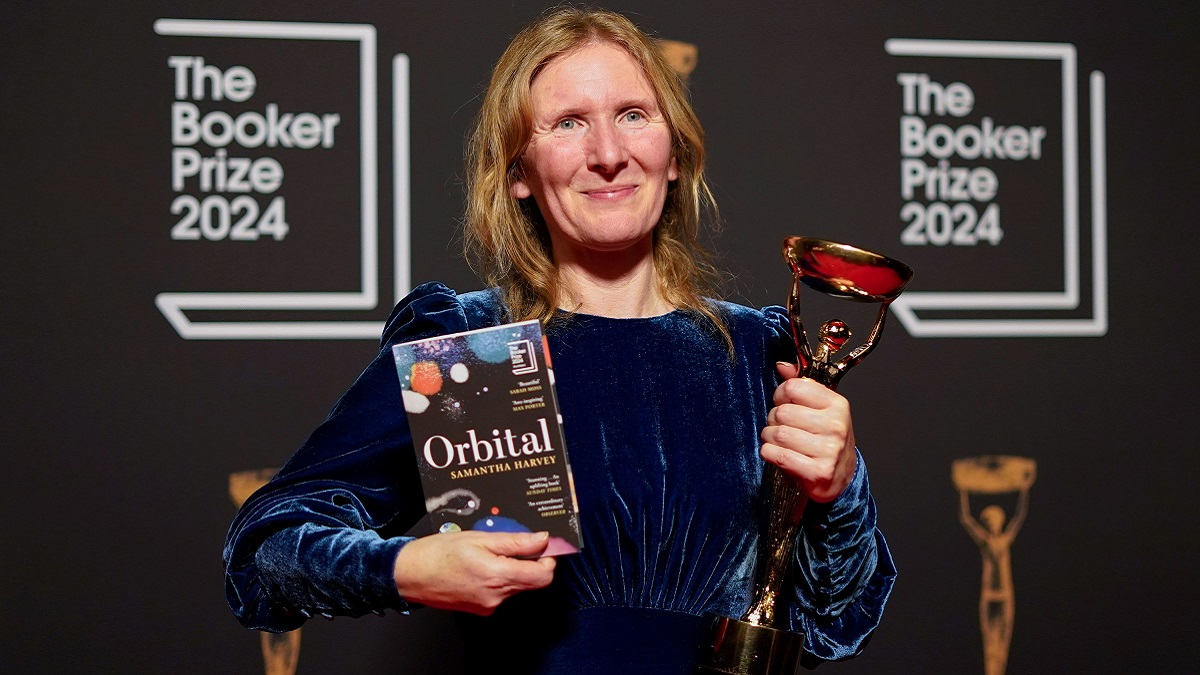 Author Samantha Harvey, wearing a dark blue dress, holds up her novel Orbital and her Booker Prize trophy.